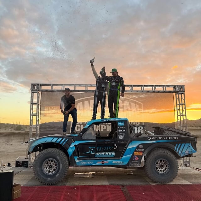 The blue Rubik Racing #18 truck on the Laughlin podium at sunset, with two people in race suits on the roof spraying champagne