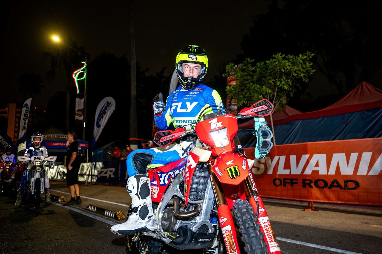 A dirt bike rider giving a thumbs up at night under a Sylvania Off Road banner at a SCORE race staging area