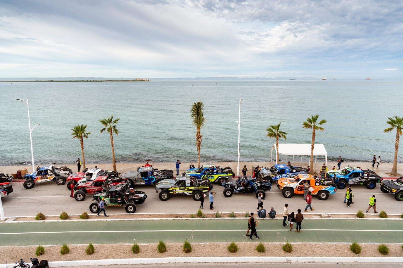 Trophy Trucks and UTVs lined up along the La Paz malecón by the water, with palm trees and spectators
