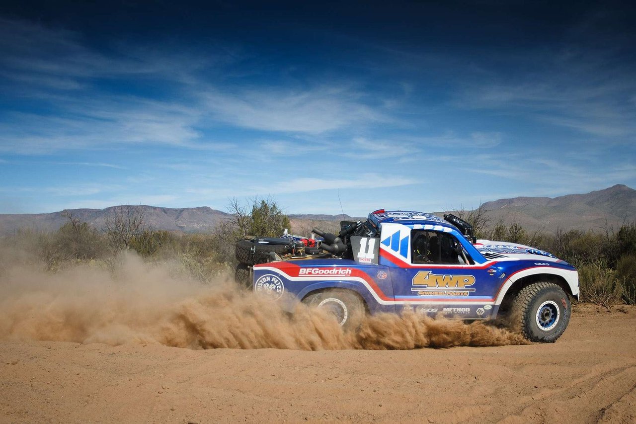 A blue, red and white No. 11 Trophy Truck kicks up a wall of dust on a desert road under a blue sky