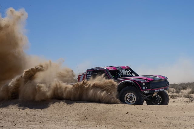A Trophy Truck throws a tall rooster tail of silt across the desert under a blue sky