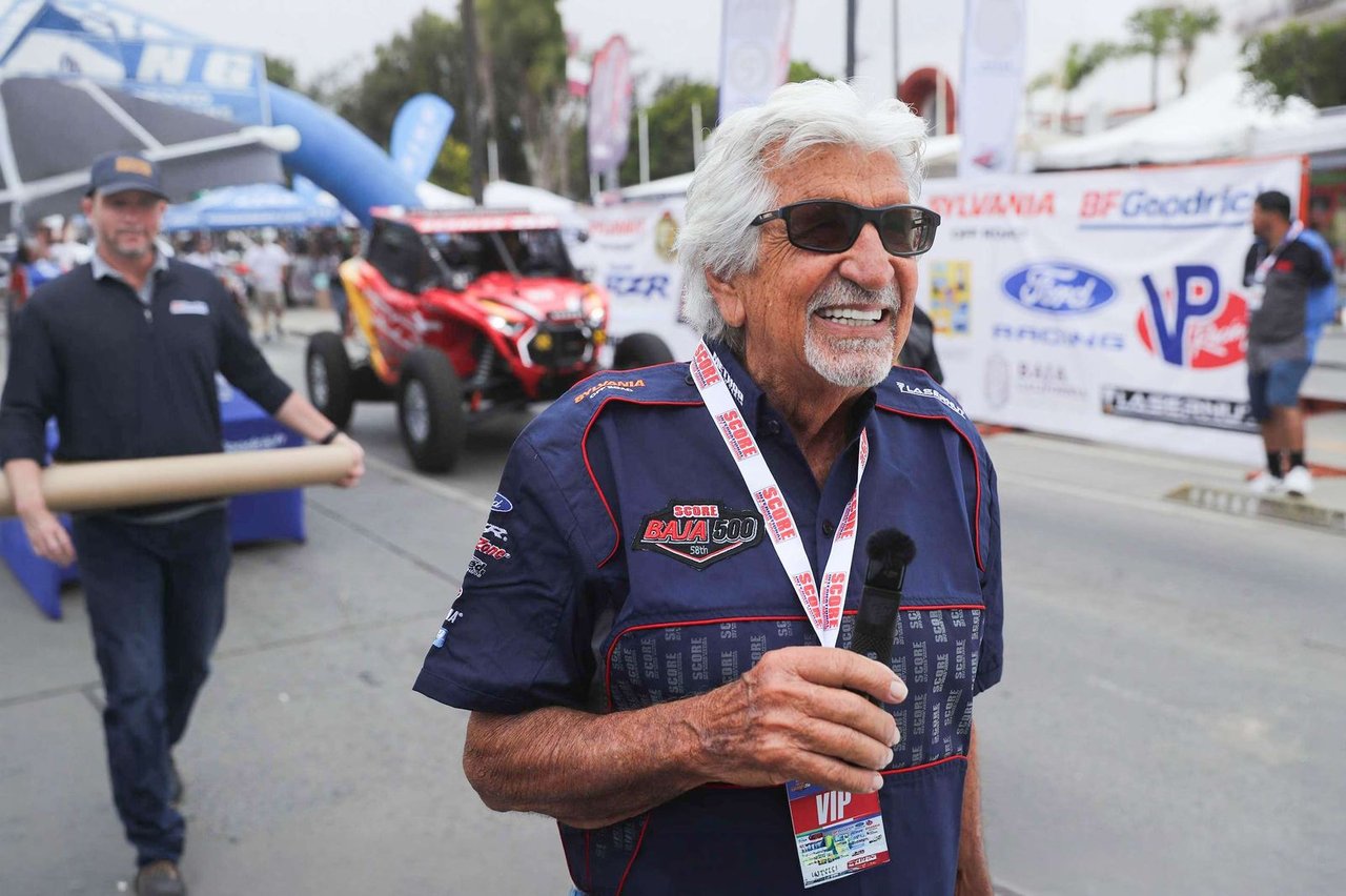 Sal Fish smiling at a SCORE race start area, wearing a Baja 500 shirt, with a red UTV and sponsor banners behind him