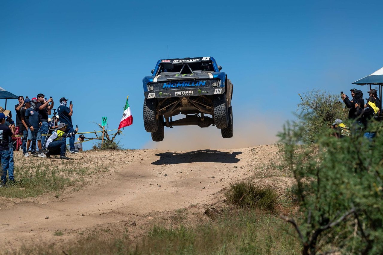 The No. 83 McMillin Trophy Truck airborne over a dirt crest as spectators along the fence raise their phones