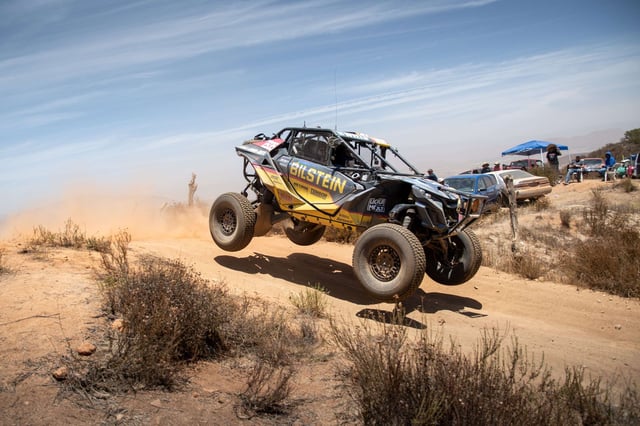 The No. 2935 Can-Am Maverick R airborne on a dusty dirt road with spectators and tents behind